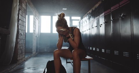 Beautiful upset sporty blonde woman sitting alone in dark gym locker room, wiping face tired after bad day slow motion.