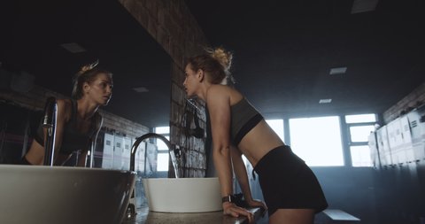 Beautiful tall sporty blonde woman washing face, looking in the mirror in dark gym locker room exhausted slow motion.