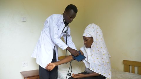Wide shot of a young male African doctor checking the blood pressure of a Muslim African lady in a small health clinic in rural Africa.
