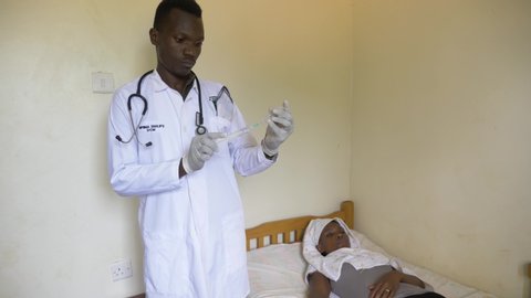 A wide shot of an African male doctor preparing an injection of medicine for a sick African woman lying in a bed at a rural health clinic.