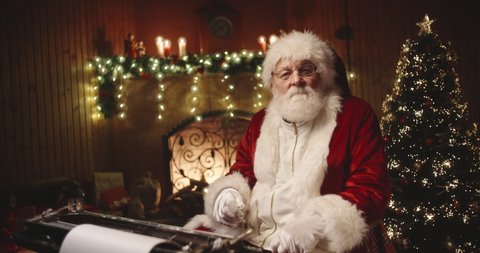Joyful santa claus sitting at his typewriter, looking at camera and playfully winking in specially decorated room - christmas spirit, holidays and celebrations concept 4k footage