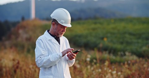 Senior technician man in protective helmet and suit using modern app smartphone working on rural field with wind turbines.