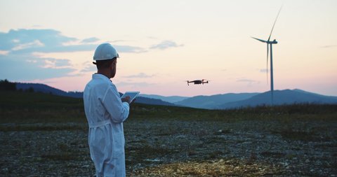 Ecology specialist in white uniform holding tablet cmputer controlling flying drone working at wind power station under sunrise. Modern technology.