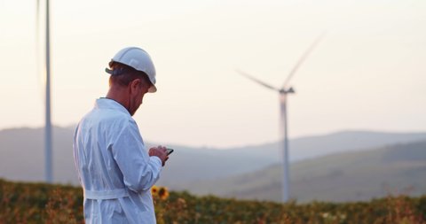 Elder engineer wearing hard hat and white costume using modern technology on smartphone working outdoor at wind turbines plant.