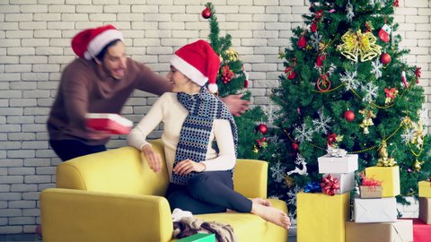 4K Wide shot playful man hugging beautiful girlfriend and surprise her a christmas gift in red box with Christmas tree background. Happy family celebration festival, love and romantic moment concept.