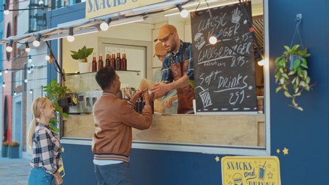 Food Truck Employee Hands Out a Burger to a Happy Young Man in Leather Jacket. Indian Man is Using Contactless Bank Debit Card to Pay for Food. Street Food Truck Selling Burgers Outdoors.