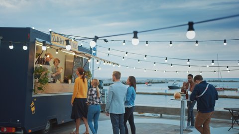 Food Truck Employee Hands Out Beef Burgers, Fries and Cold Drinks to Happy Hipster Customers. People are Eating at Tables Outside. Commercial Truck Selling Street Food in a Modern Place Near the Sea.