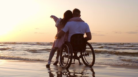 Handicapped man in wheelchair and his girlfriend on a beach at sunset
