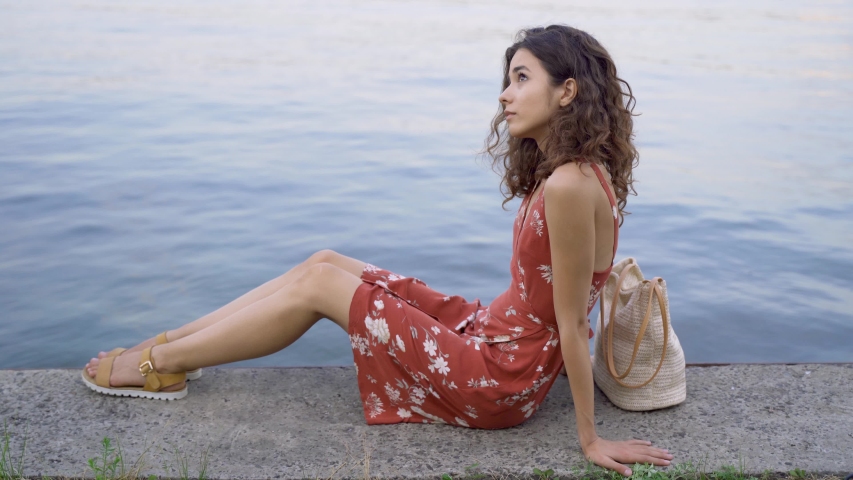 Attractive brunette with curly hair sits on a pier near the river