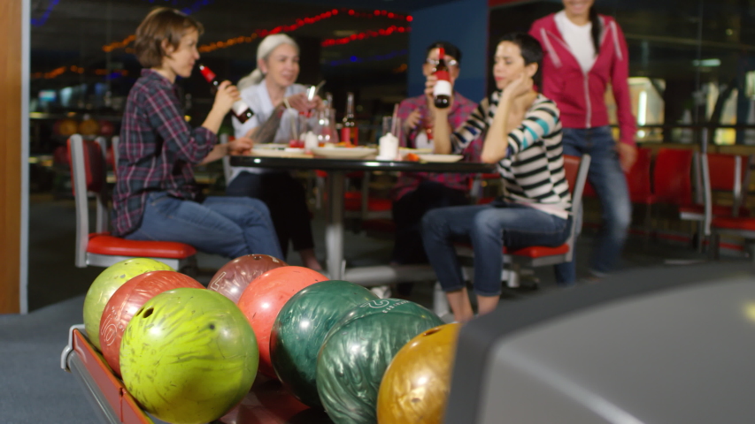 Medium shot of unrecognizable woman taking ball and knocking over pins at bowling alley as her female friends sitting at table and celebrating