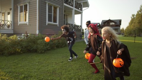 Following shot of four multiethnic teenagers wearing colorful Halloween costumes with plastic pumpkins in their hands running outdoors