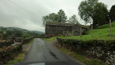 WINDERMERE, ENGLAND - 7 SEP 2018: England narrow country road hedge rows old homes POV. Historic old stone and rock homes along narrow winding roads. Hills, valley and mountain region.
