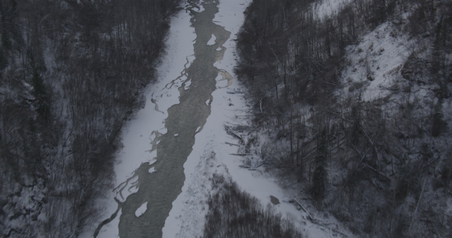Aerial helicopter shot above winter valley with dark evergreens, over partially frozen lake towards mountains, drone footage