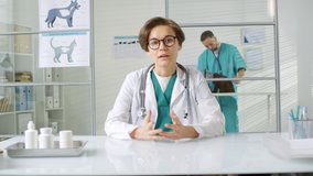 Medium shot of female veterinarian sitting at desk and having conversation with sick cat owner, while her male assistant making additional cat examination on background