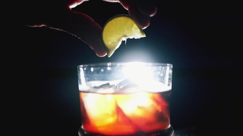 Squeezing lime or lemon onto fresh fruit cocktail. Whiskey and cola with ice cubes on dark background and studio light. Slow motion 100fps.