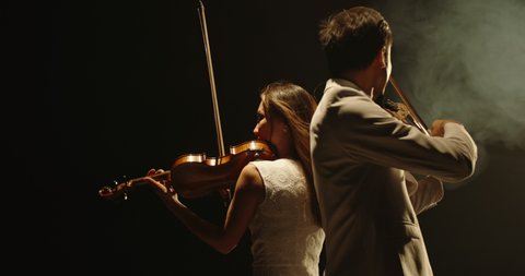 Male and female violin players having an amazing solo, standing spine-to-spine on stage, spotted by light on smoked black background 4k footage