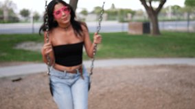A beautiful hispanic woman hipster smiling and playing and swinging on a park playground swing set with retro pink sunglasses in slow motion.