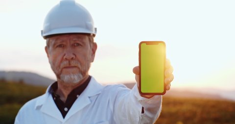 Lviv, Ukraine - September 10, 2019: Portrait of experienced technician in working suit holding mock-up smartphone green template screen staying in agricultural field.