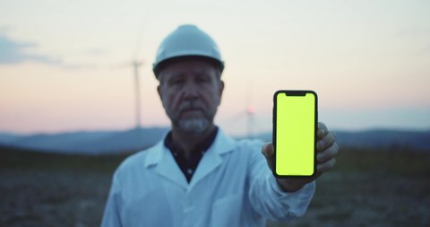 Lviv, Ukraine - September 10, 2019: Caucasian male technician holding mock-up modern smartphone technology greenscreen working at wind turbines station at sunset.
