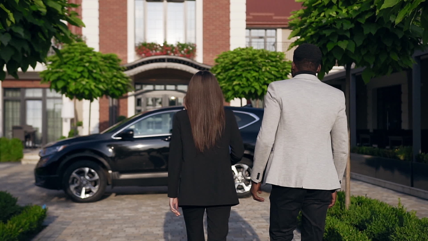 Back view of attractive two business people going to the black vehicle while driver opening the back door for their passengers and invites they comfortable in a seat. Outdoors. Slow motion