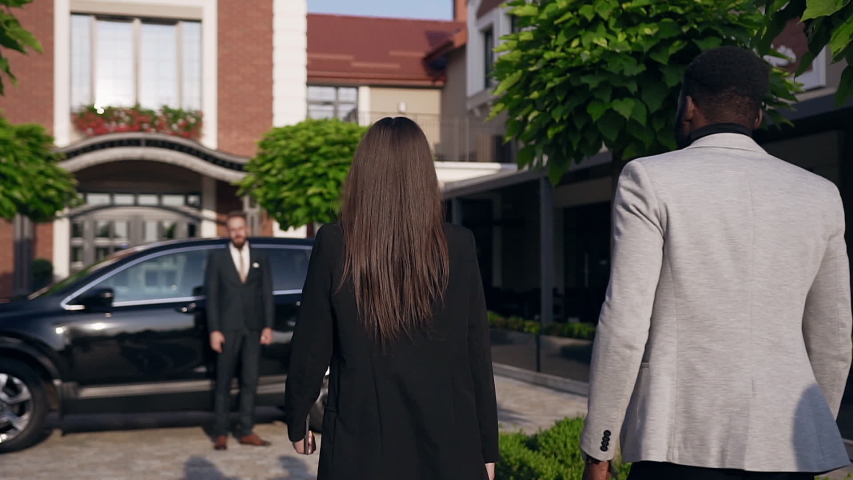 Back view of attractive two business people going to the black vehicle while driver opening the back door for their passengers and invites they comfortable in a seat. Outdoors. Slow motion