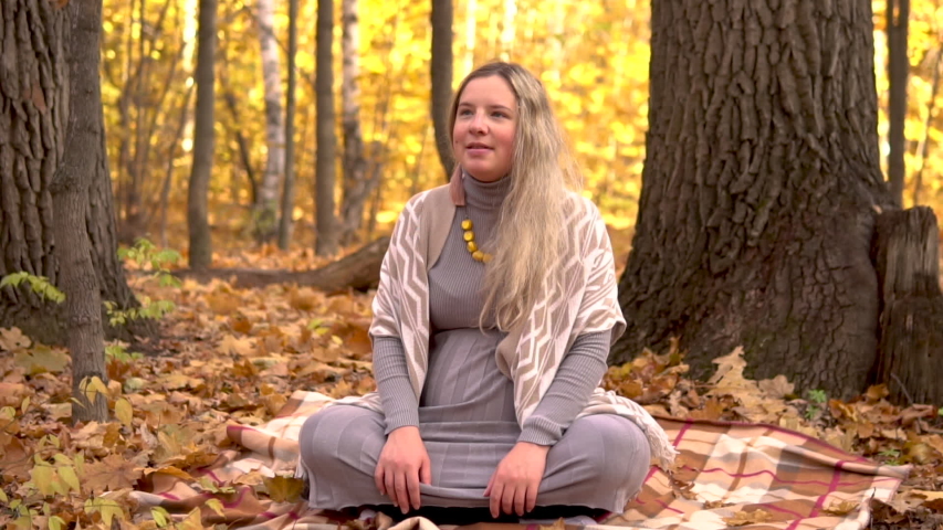 A pregnant woman is sitting under a tree in the autumn forest. Yellow leaves are falling. Slow motion