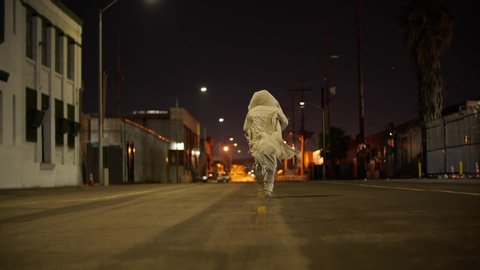 Jogging man in beige costume with hood and fluttering mantle at poor street of Los Angeles, California, USA. Night at the city, street is empty, some light from metro are on the background. Man runs