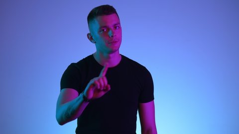 Young handsome caucasian brunette man disagrees with something looks unsatisfied. Guy shows disagreement NO gesture with his hand in studio isolated on colored background in vivid neon lights