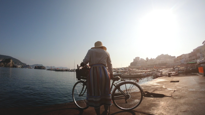Happy young woman walking with vintage bike along the harbour in Ponza island Italy. Fashion colorful dress skirt and hat. 