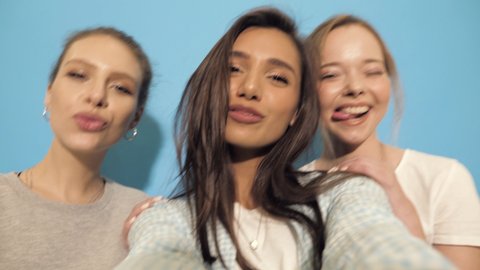 Three young smiling hipster women in summer dresses.Girls taking selfie self portrait photos or video on smartphone.Models posing in the studio near blue wall.Making duck face, show positive grimace