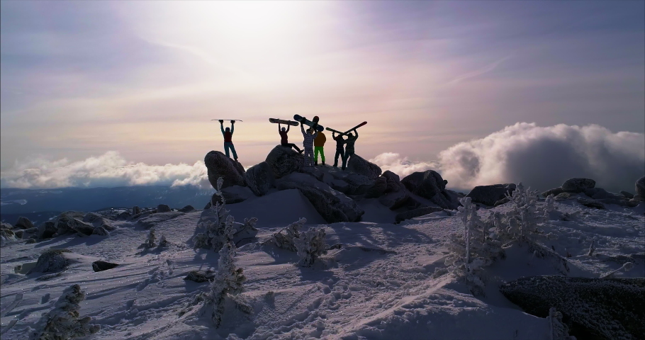 Group of happy friends skiers and snowboarders stands at sunset mountain top and having fun. Group of people stands against mountains at sunset. air camera in motion