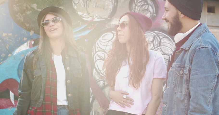 Two Caucasian beautiful and stylish young women and handsome guy, hipsters friends talking and laughing while standing at the graffity on the wall. Outdoor.