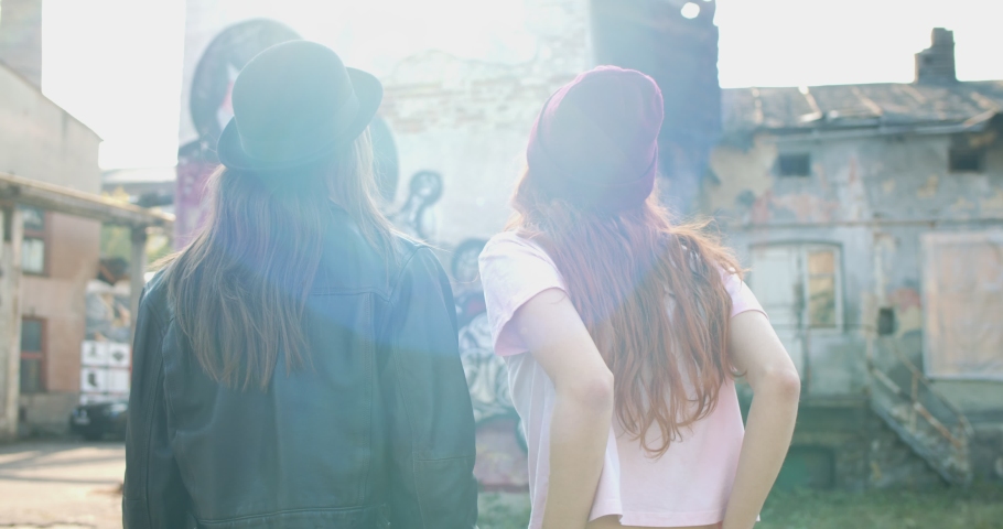 Rear of the two Caucasian young stylish hipsters friendly girls walking together and laughing at the slums with graffity on walls. Back view.
