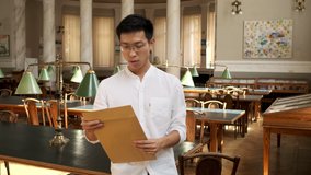 Portrait of young asian male student excitingly opening envelope with exam results in library of university