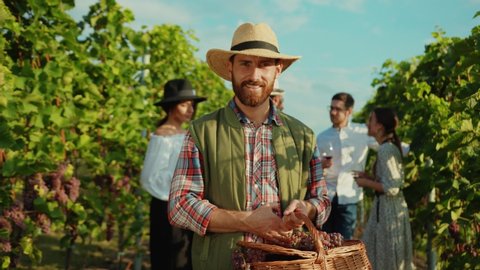 Portrait of proud caucasian farmer in strawhat smiling with joy holding large basket of grapes working during harvest in vineyard.