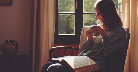 Woman Drinking Tea or Coffee and Reading a Book, Enjoying Cozy Morning. SLOW MOTION. Girl with a steaming cup of hot drink turning pages of a paper book. Cinematic Autumn aesthetics.