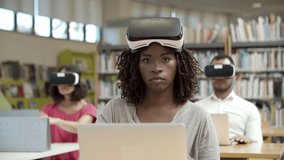 Focused African American woman wearing virtual reality glasses. Front view of excited women sitting in library and experiencing new device. Technology concept