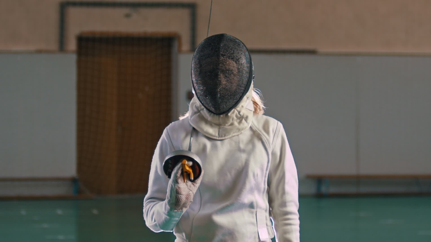 A young woman fencer with long blonde hair takes off a protective helmet holding a sword | Shutterstock HD Video #1038494981