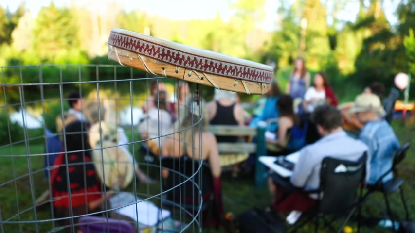Sacred drums at spiritual singing group. Blurred people are seen in a singing circle at a local park during a sunny afternoon, details on the side of a handmade native drum are seen in the foreground.