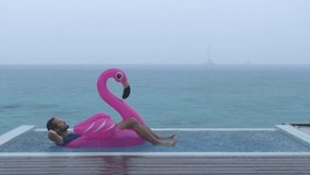 Vacation rain - funny video of holidays getaway travel raining away under heavy rainfall with relaxing and happy man smiling on flamingo float in luxury pool with funny face expression.