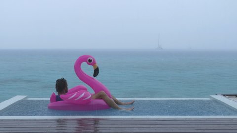 Vacation rain - funny video of holidays getaway travel raining away under heavy rainfall with sad and unhappy man on flamingo float in luxury pool with funny face expression.