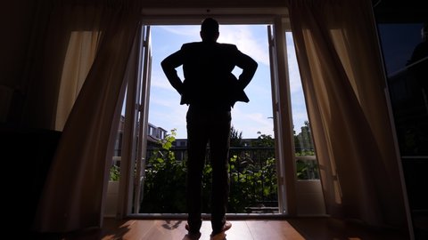 Business man unveil curtains and stay against big open window, with arms on hips. Silhouetted full length shot from back. Typical European town-house bedroom, nice sunny morning hours