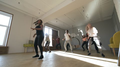 Dolly-in shot with wide angle of black female instructor and diverse group of young people rehearsing dance routine in airy studioの動画素材