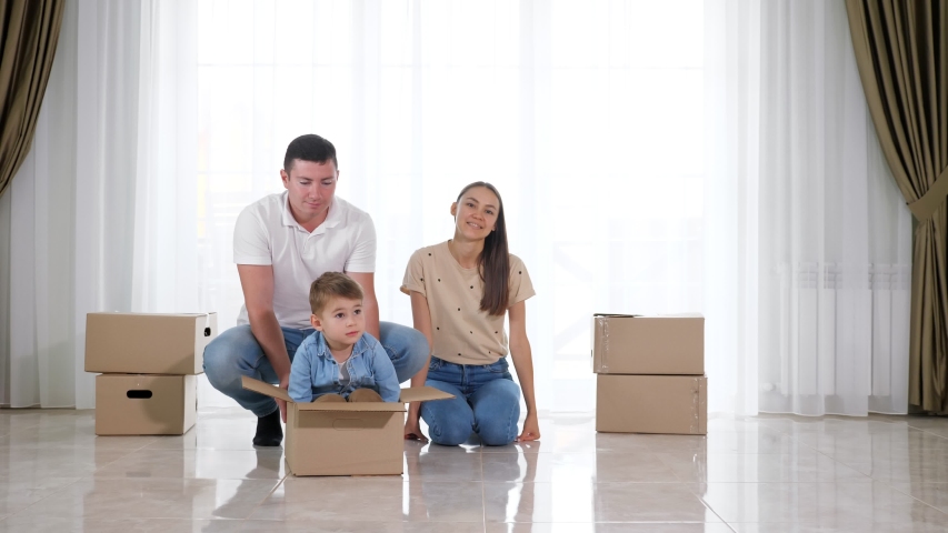 Father rides happy small son wearing blue shirt in big cardboard box near beautiful mother sitting in floor slow motion | Shutterstock HD Video #1038506489