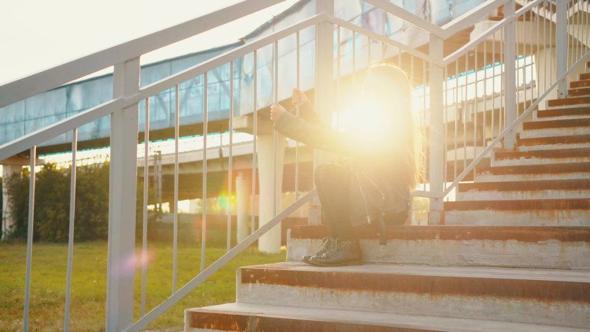 Excited stylish teenage girl posing on stairs during sunset. Low angle view of adorable girl in black leather jacket holding railing and posing on steps during sunset. Fashion concept