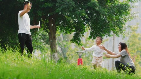 Slow motion happy Chinese family playing outdoor in summer park little Asian boy chasing soap bubble together with parents young couple having fun with son lovely mom dad wellbeing lifestyles nature