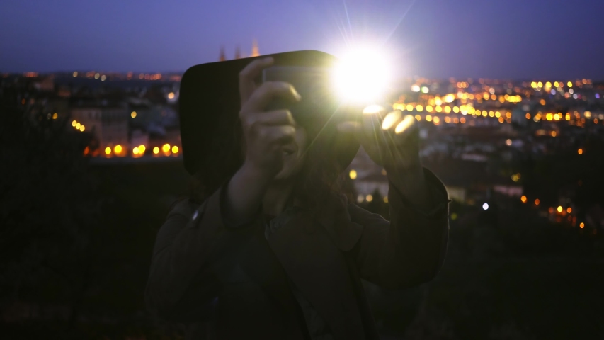 Woman using smartphone at night. Young girl in hat standing on top of hill above cityscape of Prague, she taking photo and flashing light on smartphone at blurred background illuminated city 