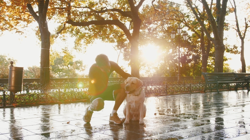 Portrait of young black man and his white labrador dog in city park during beautiful autumn morning