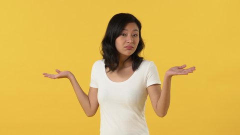 Asian girl expressing no and don't care gestures by shrugging shoulders with open hands and moving index finger isolated on yellow backgrond