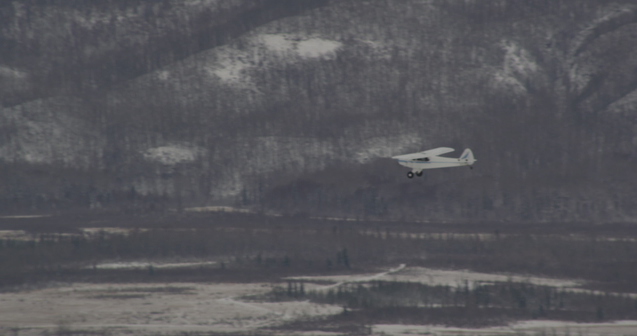 Aerial helicopter shot, small blue and white airplane flying over vast Alaskan mountain valley, banks left and flies towards camera, past it, drone footage